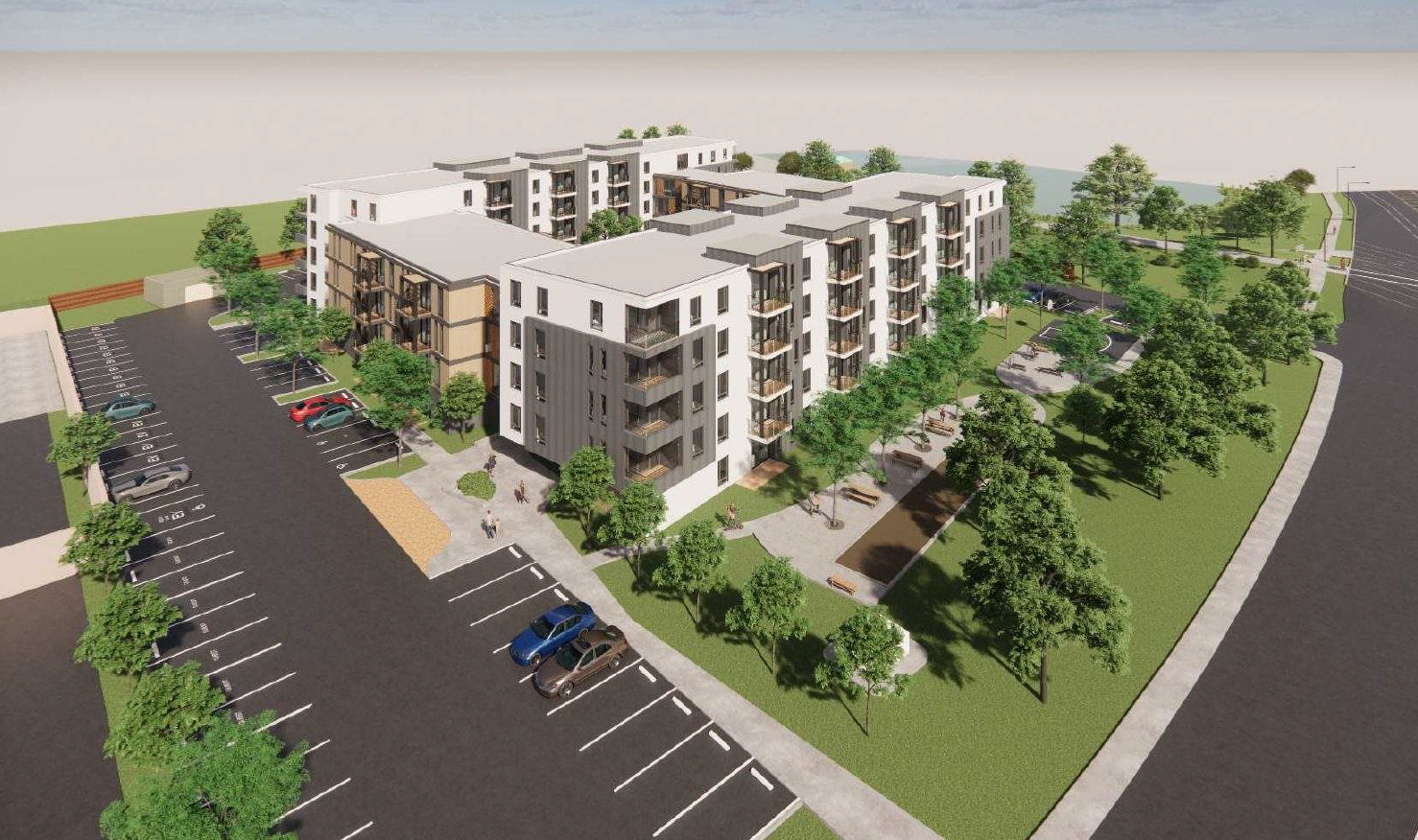 Aerial view of a modern apartment complex with multiple buildings, surrounded by green lawns and trees. A parking lot with cars is visible on the left, and pathways lead through landscaped areas in the center.