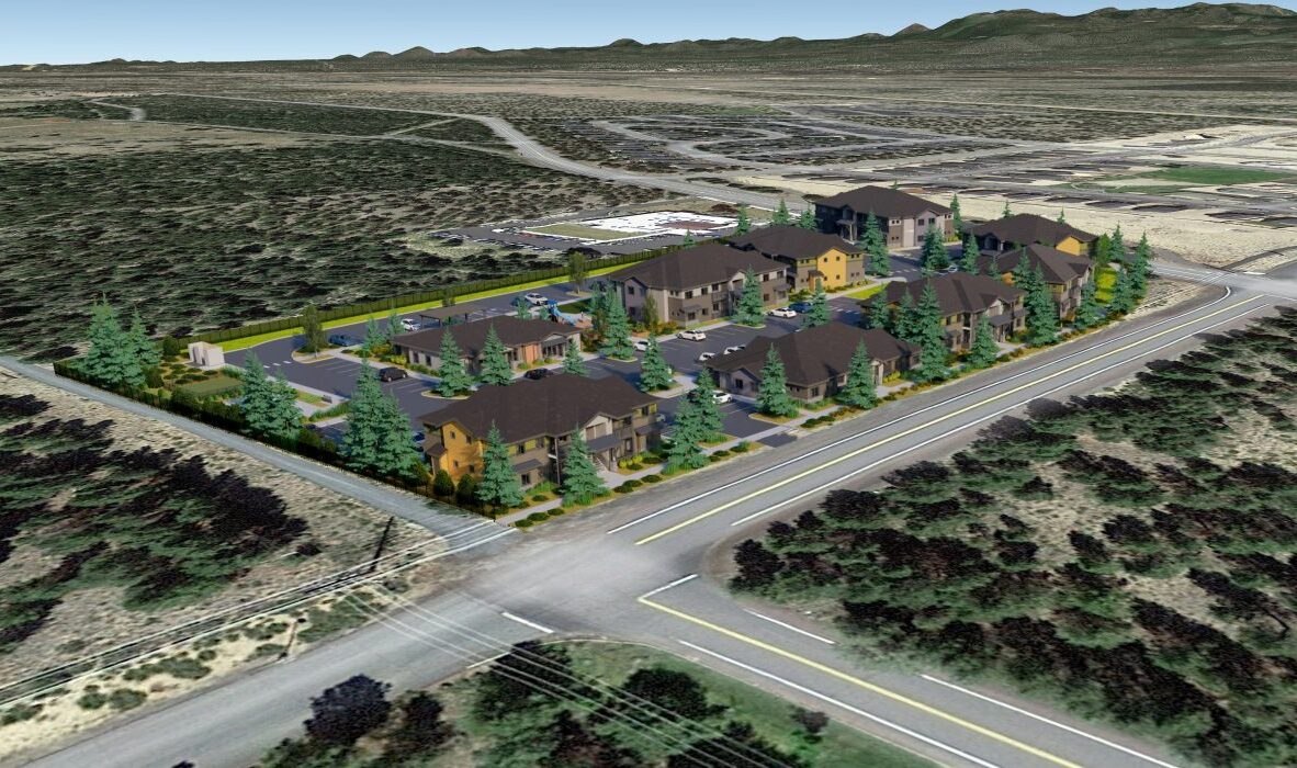 Aerial view of a housing development surrounded by desert landscape. The complex includes multiple buildings amidst green trees, situated at a road intersection. The area is bordered by open land and distant mountains.