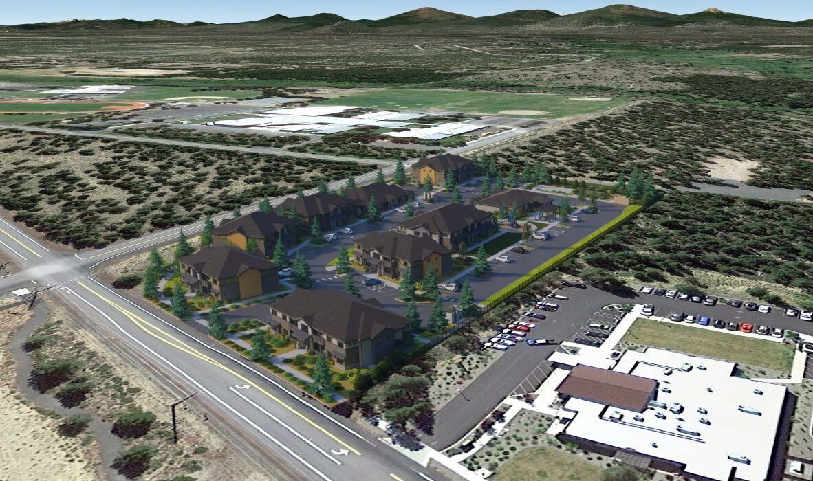 Aerial view of a residential neighborhood with several two-story buildings surrounded by trees and greenery. A parking lot is adjacent, and a larger building with a filled parking area is nearby. The landscape is partly desert with distant hills.