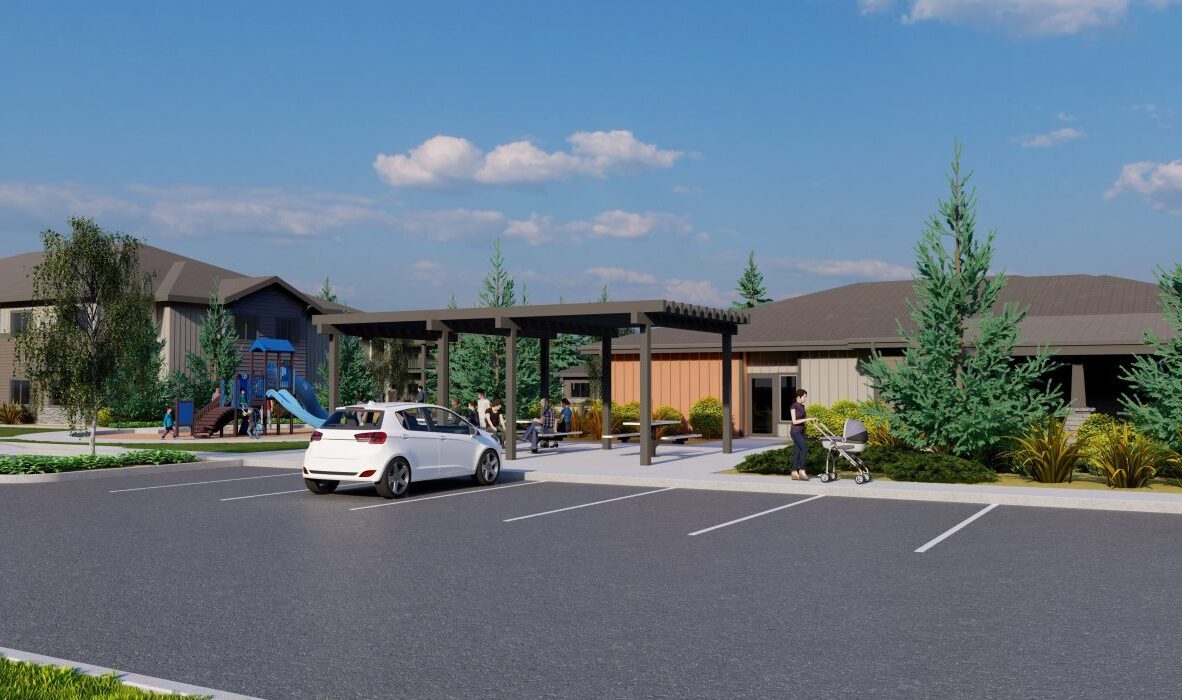 A modern residential building with a covered seating area and a playground nearby. Several people are gathered under the pergola, and a parked white car is visible. Trees and shrubs surround the parking lot. The sky is clear and blue.