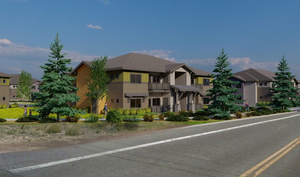 A modern two-story house with a sloped roof, surrounded by neatly trimmed grass and several tall pine trees. The road in the foreground leads to the entrance, and there are other similar houses in the background. The sky is clear and blue.