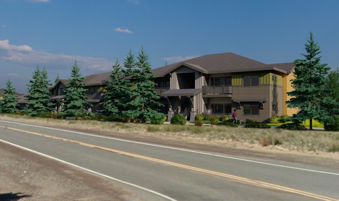 A two-story apartment building with a modern design is surrounded by evergreen trees. The building features balconies and a sloped roof. A road runs along the foreground under a clear blue sky.