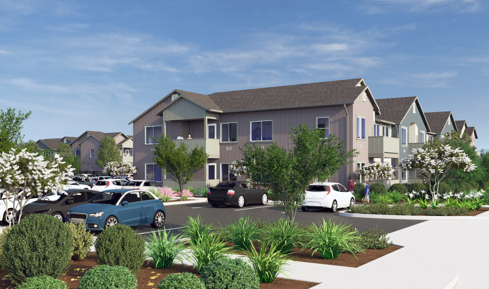A residential complex with modern two-story buildings surrounded by trees and shrubs. Several cars are parked in the lot, and people are walking on the sidewalks. The sky is clear and blue.