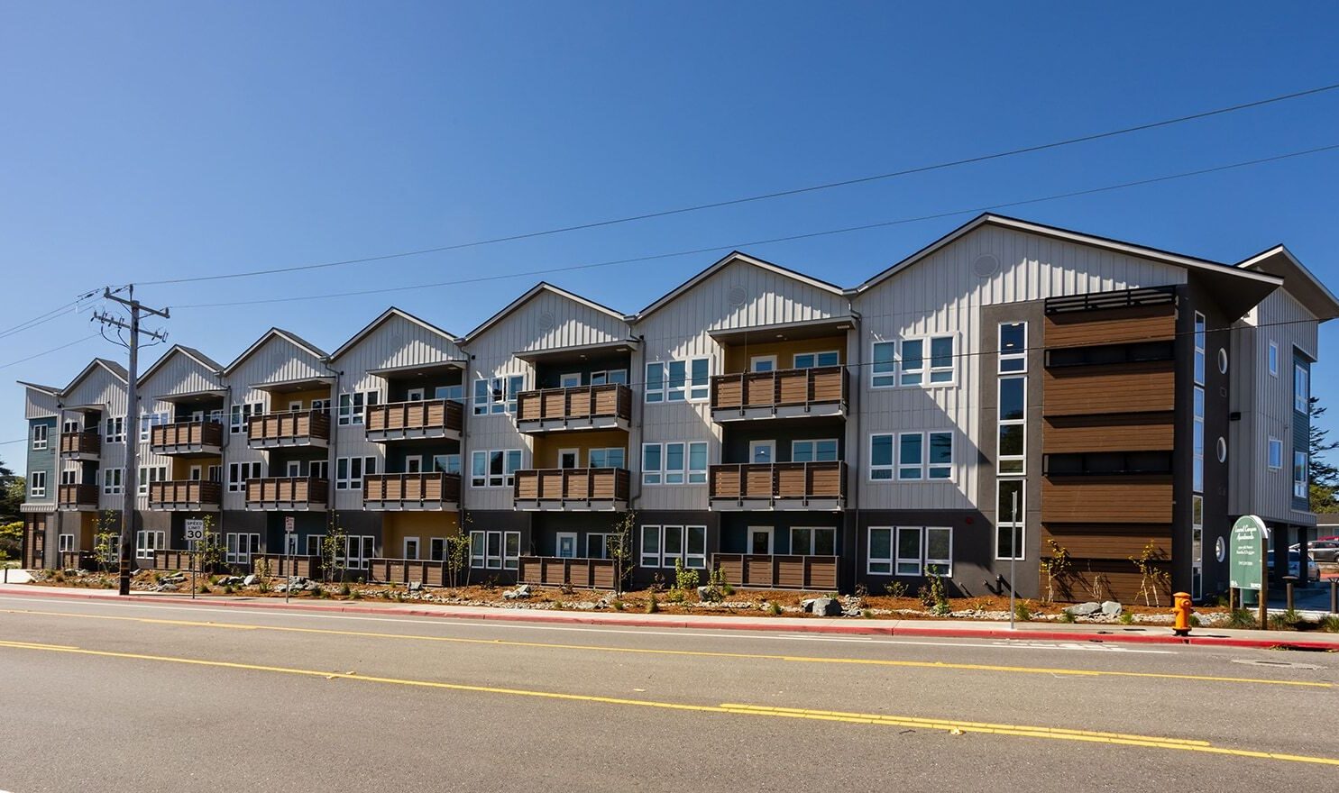 A modern three-story apartment building with a gray and brown facade, featuring balconies on each level. The building is set along a street with power lines and clear blue skies in the background.