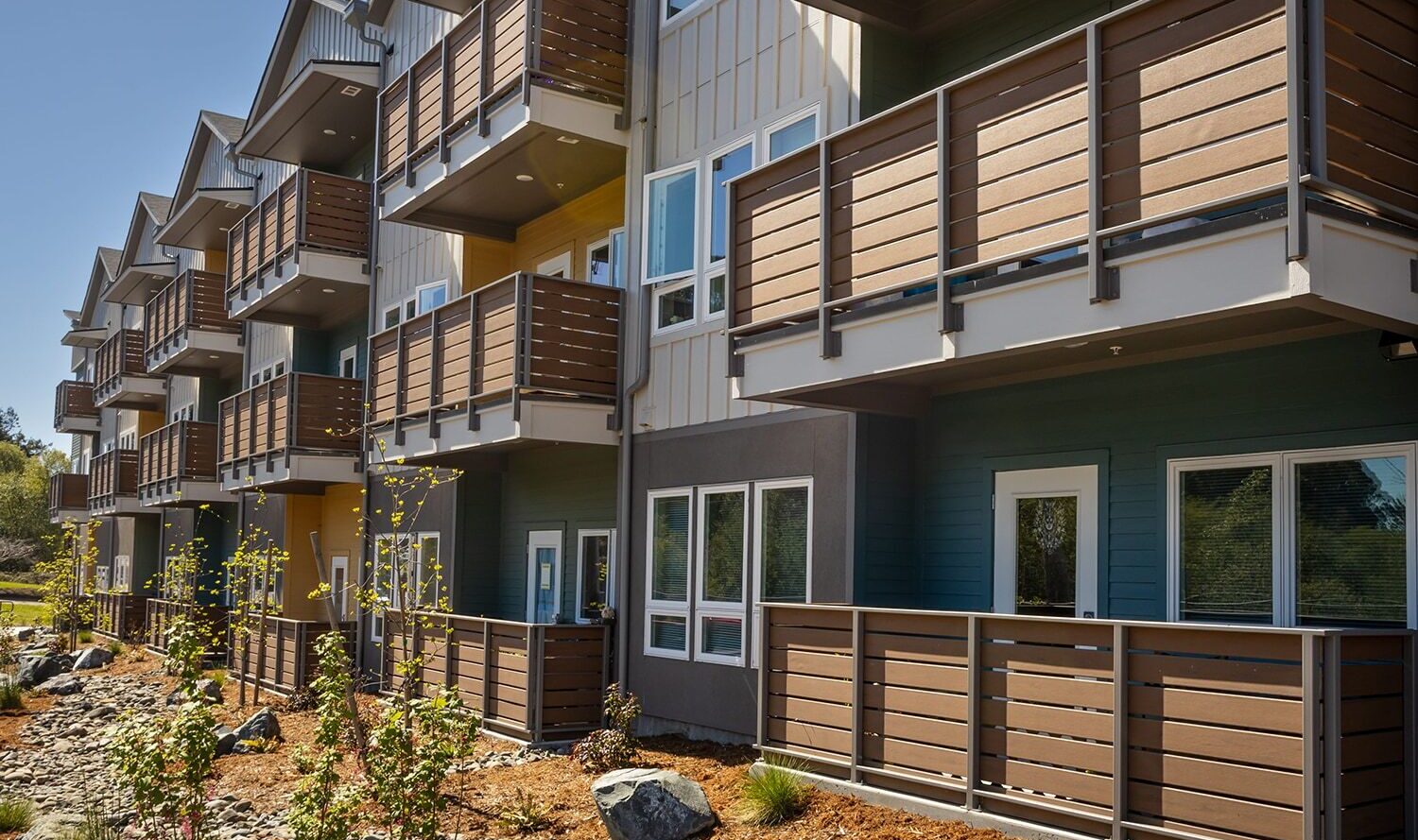Modern apartment building with several floors and wooden balconies. The architecture features clean lines and a mix of brown and beige colors. Young trees are planted along the landscaped garden bed in front.