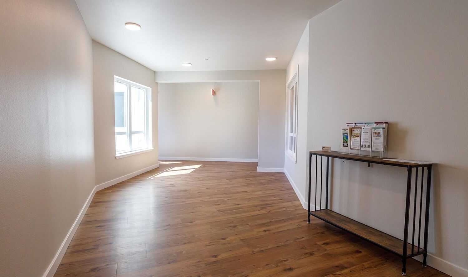 An empty room with light wooden flooring, white walls, and a large window letting in natural light. A narrow console table with a few brochures is against the right wall. The room has recessed lighting on the ceiling.