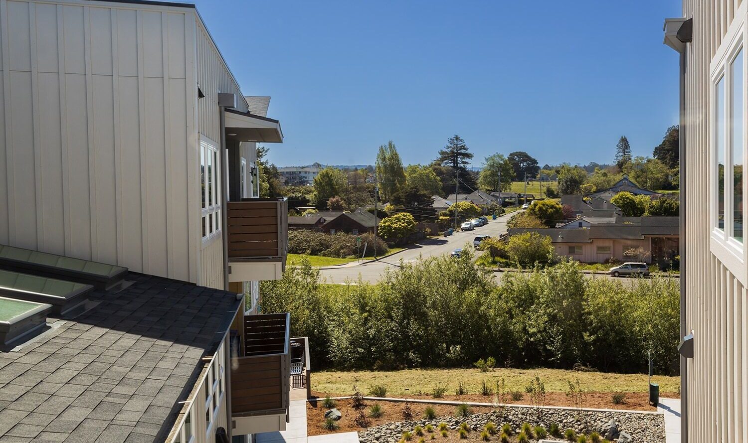 View from a building overlooking a suburban neighborhood with houses, trees, and a road. There is a landscaped garden in the foreground and clear blue sky in the background.