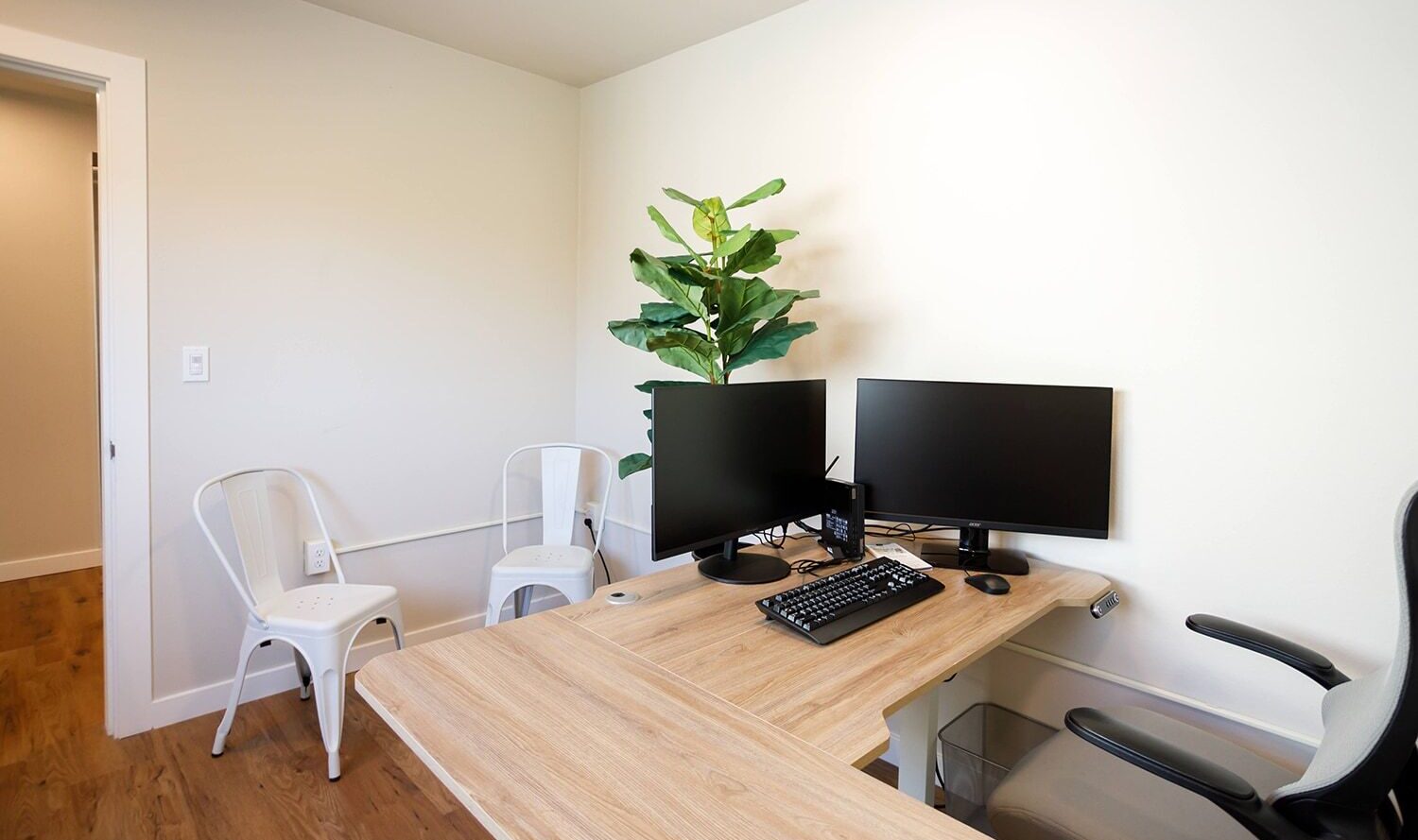 A minimalist office setup featuring a wooden desk with a keyboard, two monitors, and a potted plant. There are metal and fabric chairs around the desk, and the room has wooden flooring and white walls.