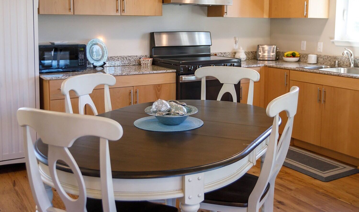 A cozy kitchen with wooden cabinets and granite countertops features a round table with four white chairs. The table is set with a blue plate and a decorative bowl. Track lighting overhead and a window provide natural light.