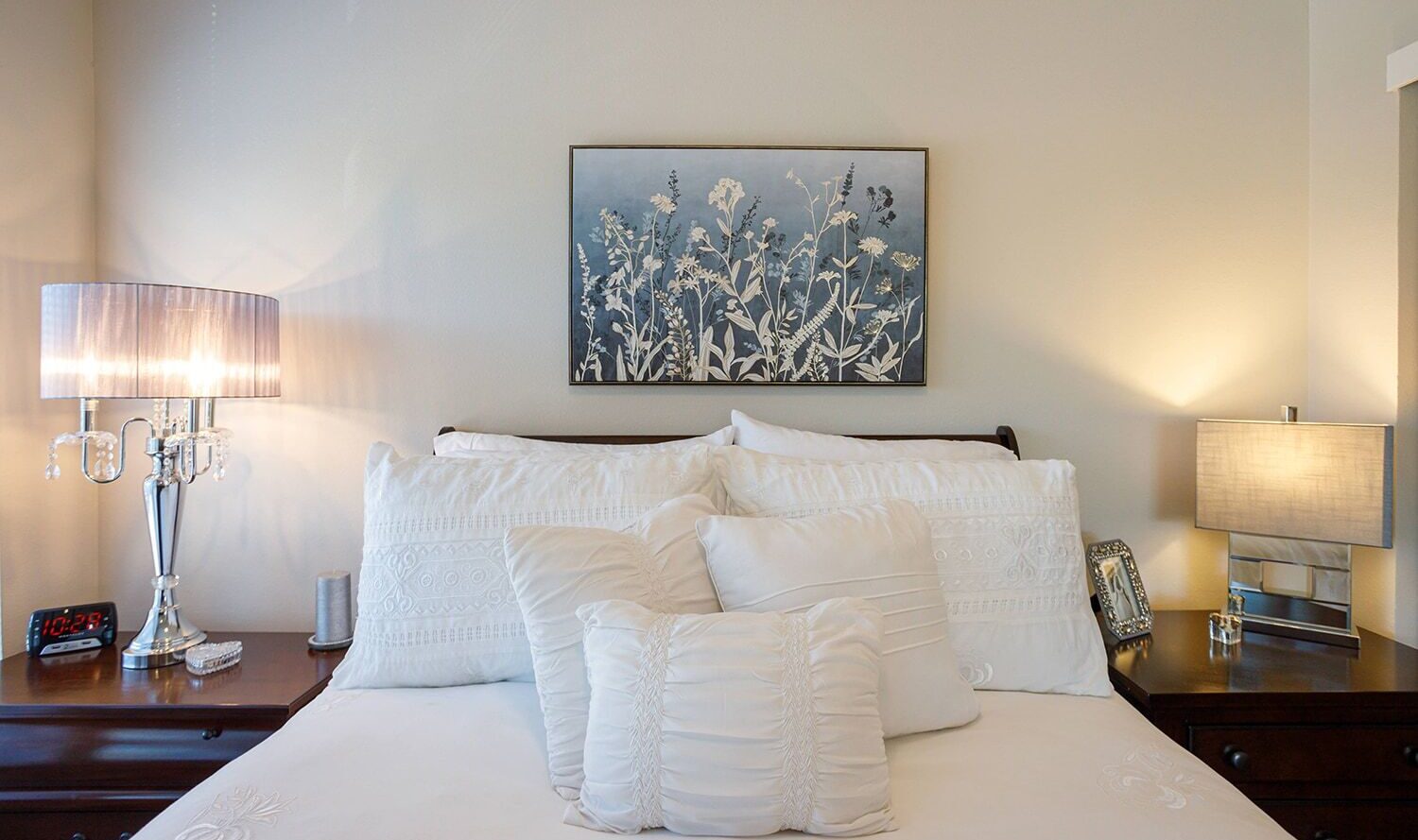 A neatly arranged bed with multiple white pillows and embroidered linens. Two bedside tables each have a lit lamp and a digital clock displaying 12:52. A framed floral artwork hangs above the bed against a light-colored wall.