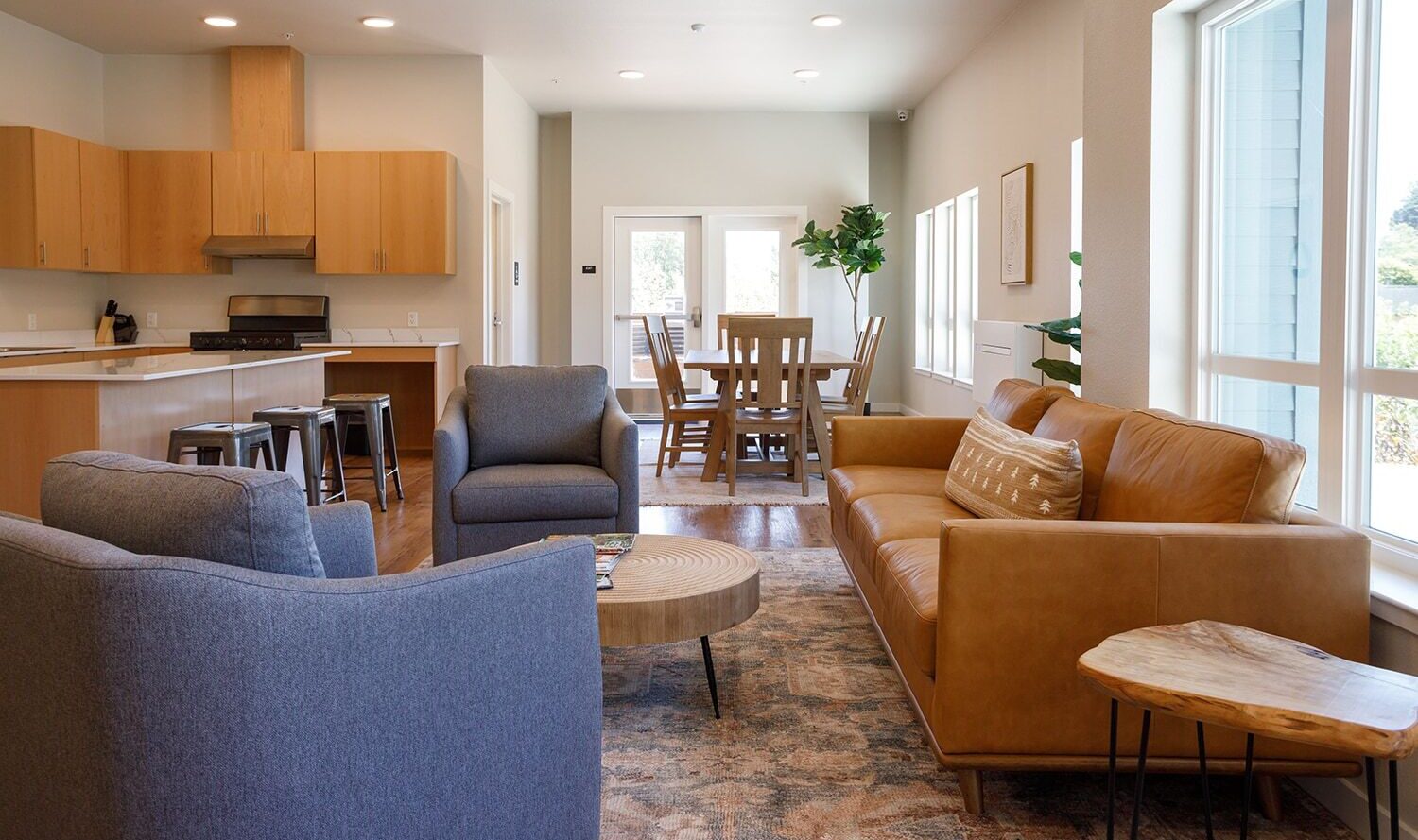 A modern open-concept living and dining area featuring a tan leather sofa, two gray armchairs, a wooden coffee table, and a dining table with chairs in the background. The room has natural light from large windows and wooden cabinetry in the kitchen area.