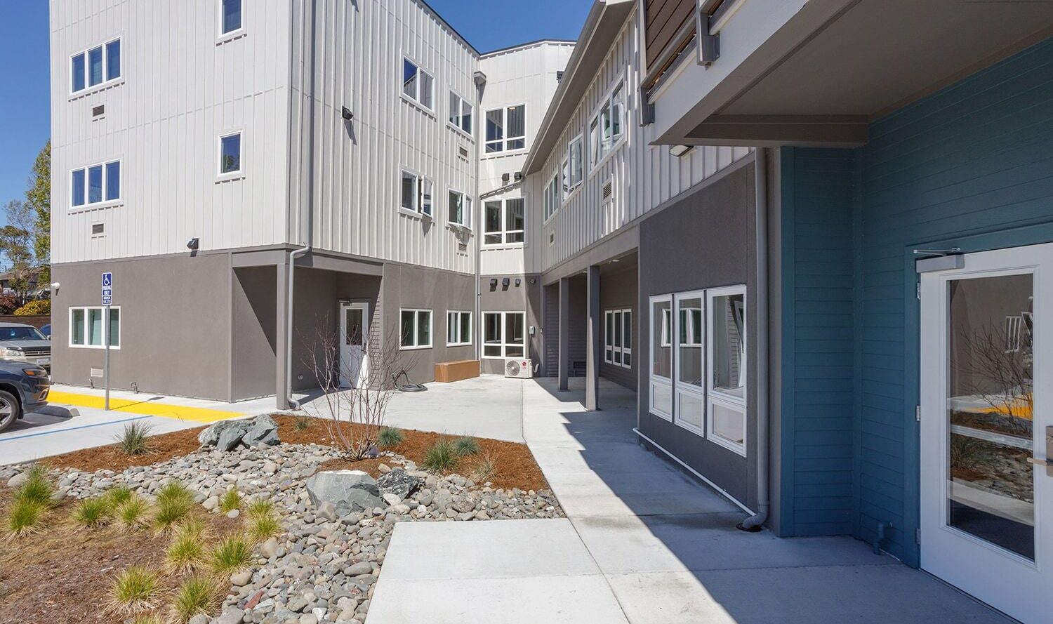 Modern, two-story building with gray and white siding. The exterior features multiple windows, a small landscaped area with rocks and plants, and a clear entry path. The sky is clear and blue.