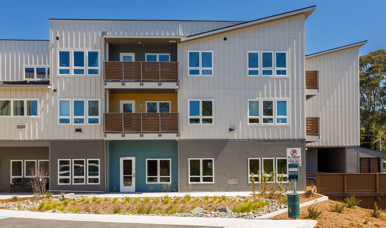 A modern, three-story apartment building with a gray and blue facade. Large windows and brown balconies accentuate the design. The front entrance is surrounded by a small landscaped area and a paved walkway. Bright blue sky overhead.