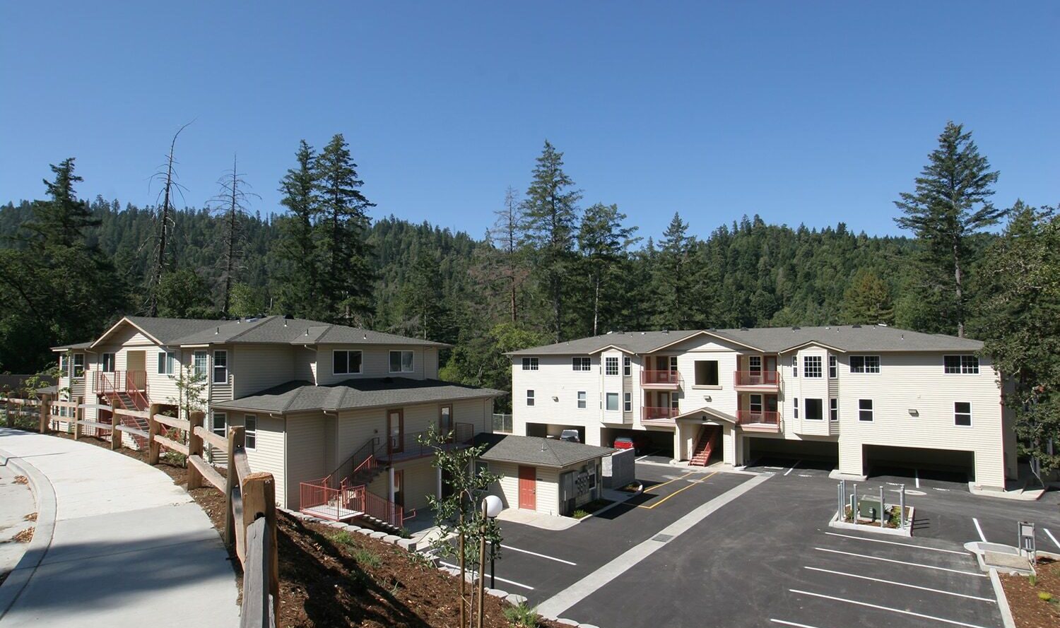 A small residential complex in a forested area featuring three beige two-story buildings with green roofs. The complex includes a parking lot, and the surrounding landscape is lush with tall trees and greenery under a clear blue sky.