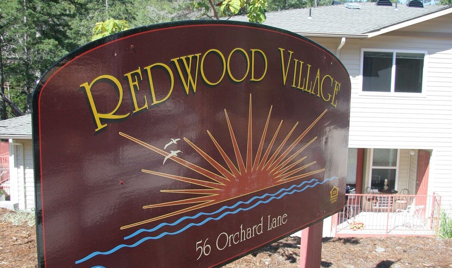 Sign for Redwood Village at 56 Orchard Lane, featuring a sunburst and a stylized river. Theres an accessibility icon on the sign. Trees and a light-colored building are visible in the background.