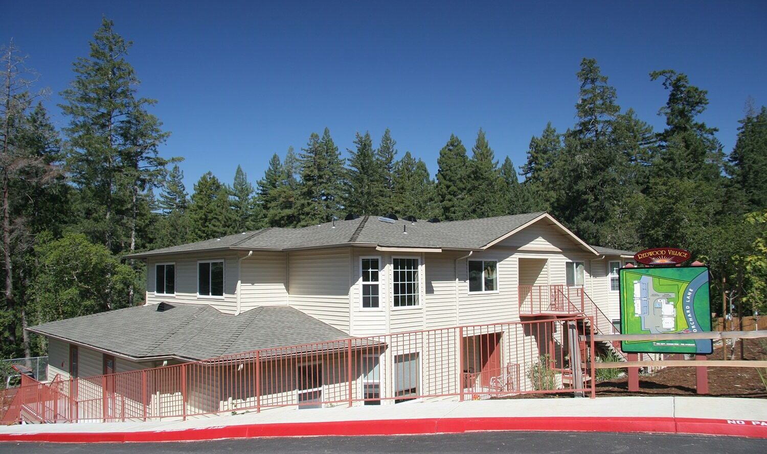 A two-story beige building with a gray roof, surrounded by tall trees. A colorful sign with a map is visible at the entrance, and a red curb outlines the parking area in the foreground.