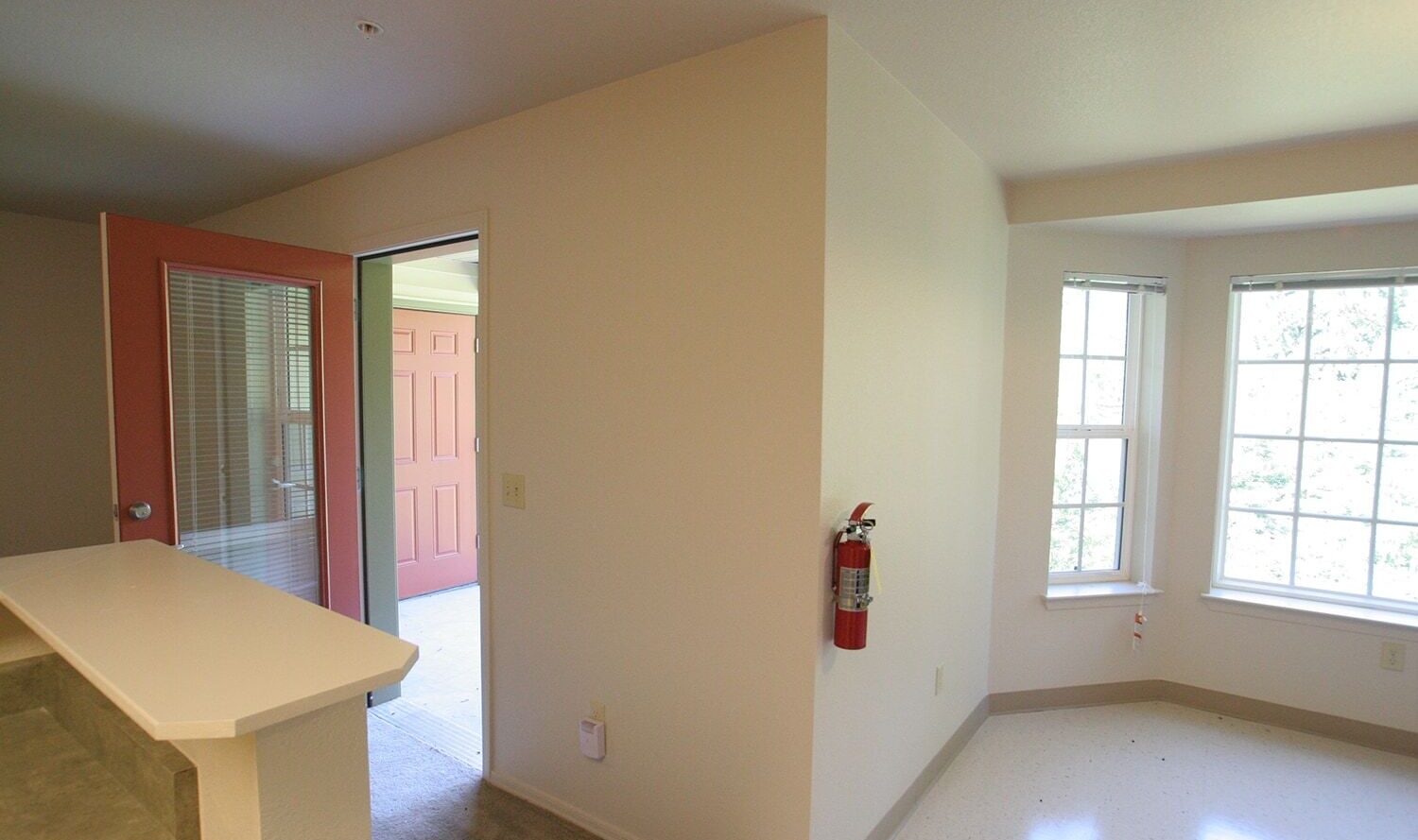 An empty room with beige walls, a fire extinguisher mounted on the wall, and large windows letting in natural light. The room opens to a small patio through a glass door with a screen. A countertop is partially visible in the foreground.