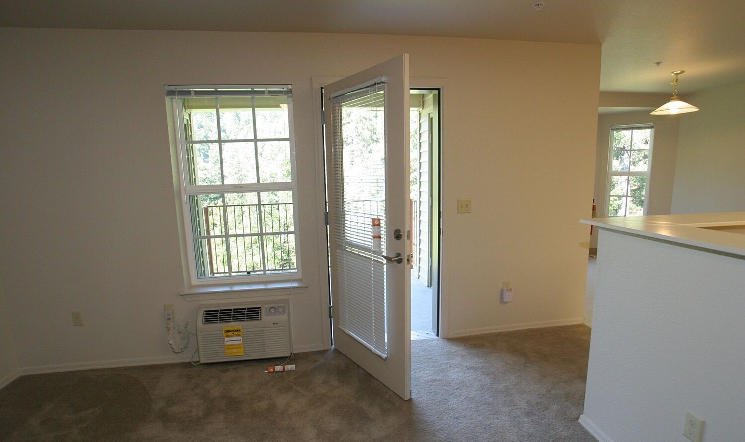 A room with beige carpet and cream walls, featuring a window, an open door leading to a balcony, a wall-mounted air conditioner below the window, and a small light fixture above a kitchen counter. Natural light fills the space.