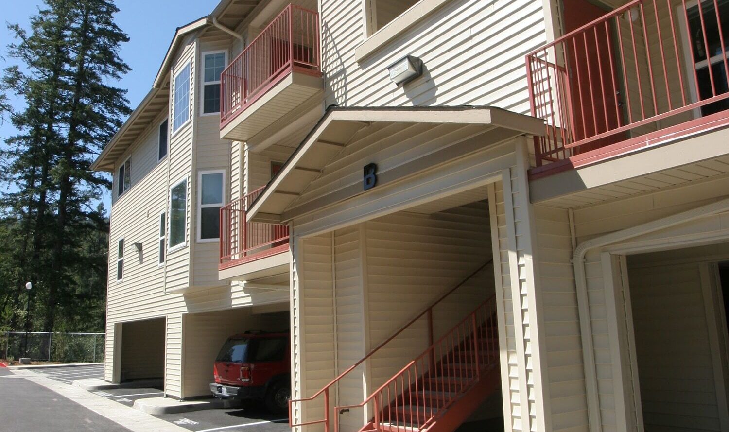 Three-story beige apartment building with red railings and a staircase. Open garages and a red car are visible in the lower level. The area is well-lit with a clear blue sky and surrounded by trees.