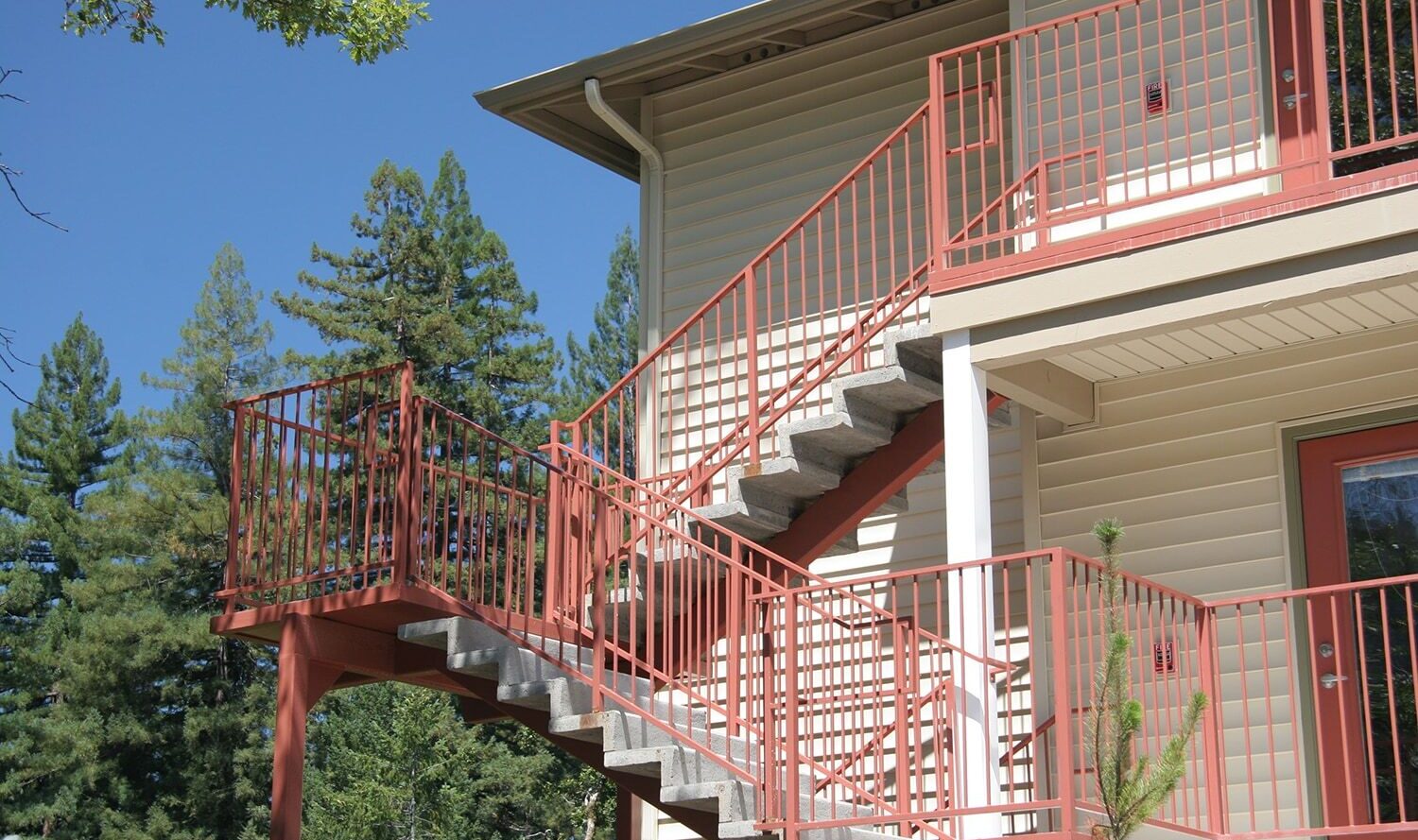 A beige building with a red metal staircase leads to the upper floor. Surrounded by tall green trees, the scene is set against a bright blue sky. The staircase includes railings and a landing area.