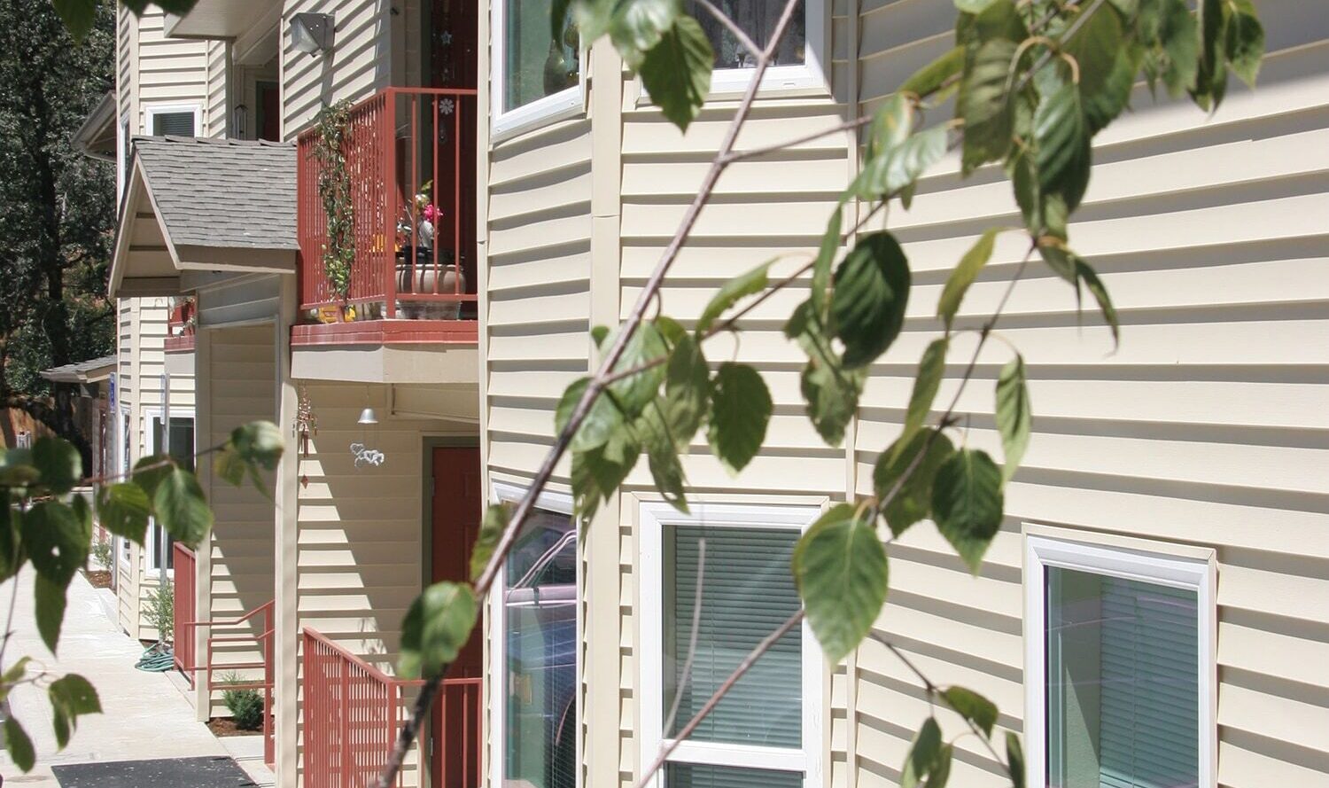 Exterior view of a beige apartment building with red balconies. A branch with green leaves is in the foreground, and there is a concrete path leading to the entrance.