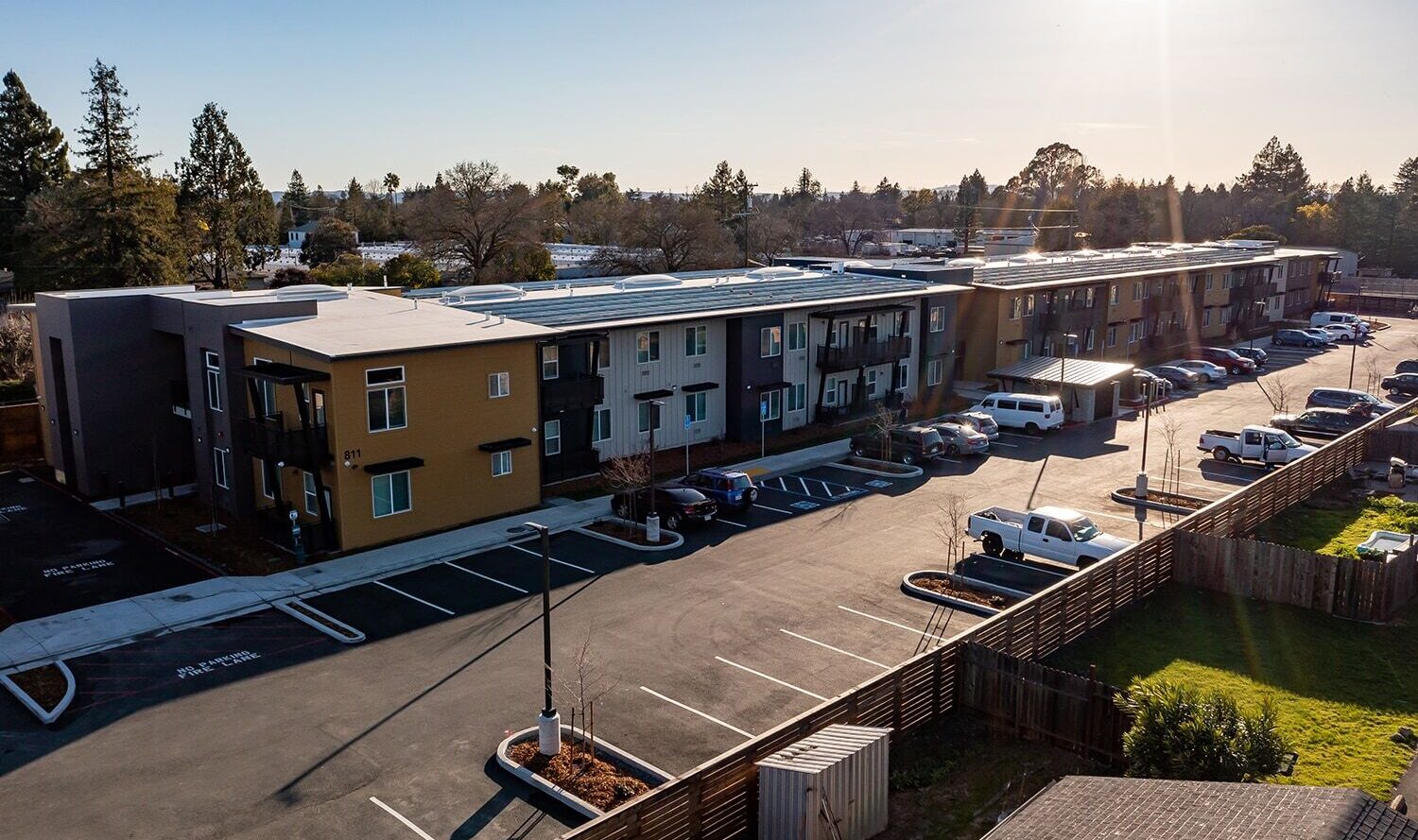Aerial view of a modern apartment complex with a parking lot full of cars. The buildings are two stories high, with a mix of brown and gray colors. Trees and greenery are visible in the background under a clear sky.