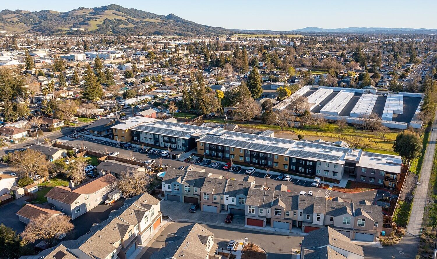 Aerial view of a suburban neighborhood featuring rows of houses, a large, colorful building in the middle, and fields with mountain ranges in the distant background under a clear blue sky.