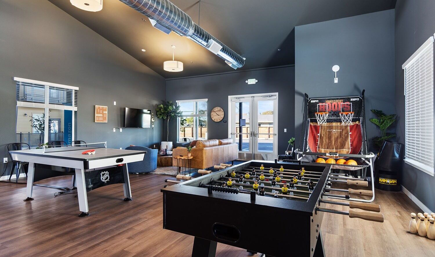 A spacious game room with gray walls featuring air hockey and foosball tables in the foreground. In the background, theres a basketball arcade machine, a television, and a seating area with couches. Large windows let in natural light.