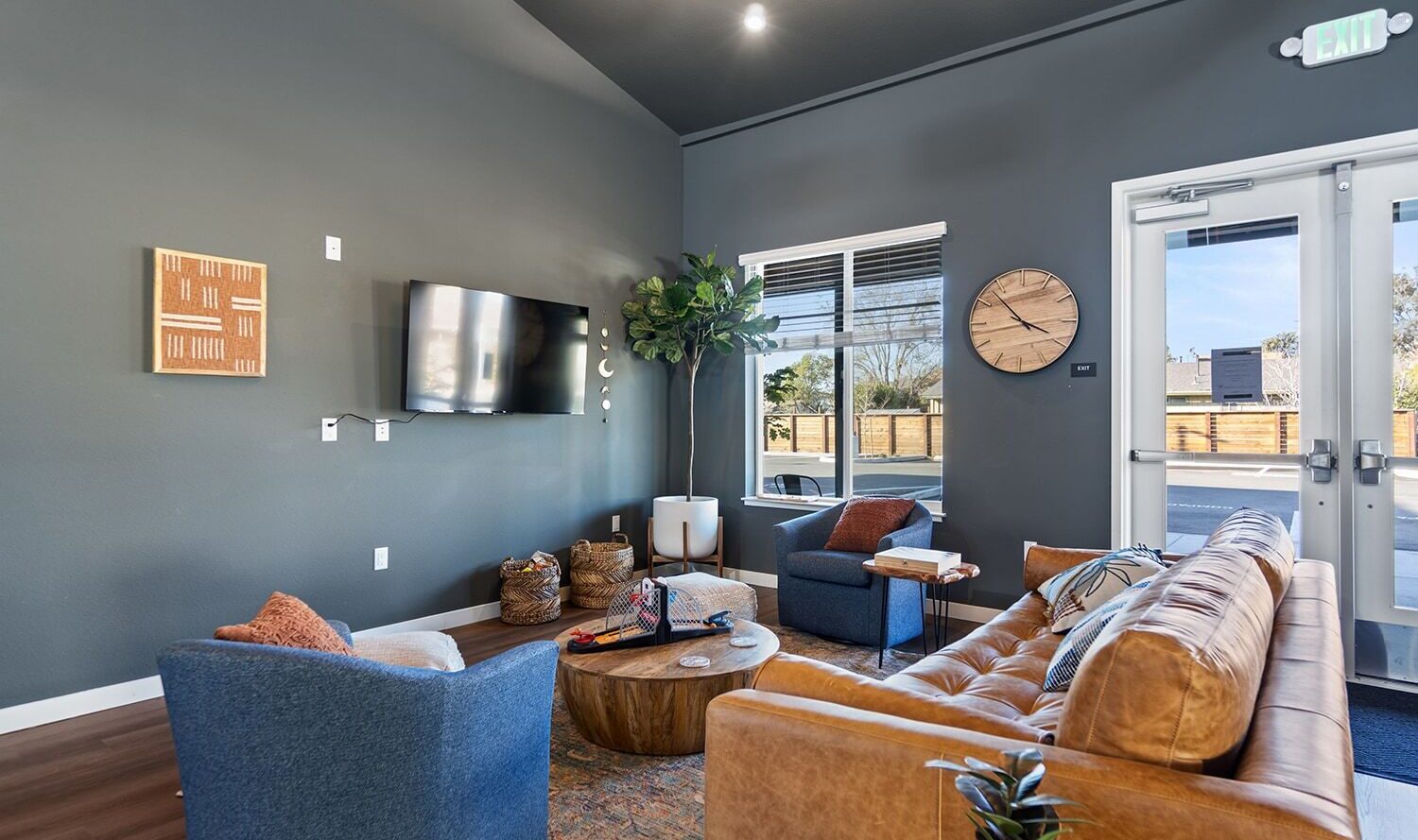 A modern lounge with a leather sofa, two blue armchairs, and a wooden coffee table. A large wall-mounted TV is on the dark wall, accompanied by a clock and artwork. A potted tree stands by a window and a glass exit door. Cozy, contemporary decor.