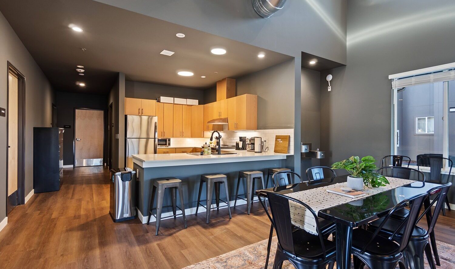 A modern kitchen and dining area with wooden cabinets, a large white island with barstools, and a black dining table with chairs. The space features hardwood floors, grey walls, and industrial-style ductwork. A potted plant sits on the table.
