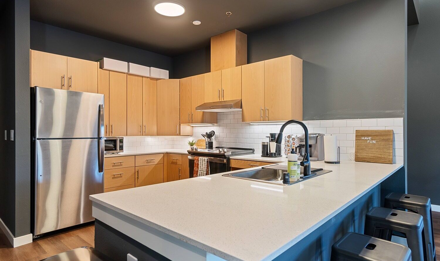 A modern kitchen with light wood cabinets, a stainless steel refrigerator, and a white subway tile backsplash. The countertop features a sink, and there are metal barstools along the island. A small sign reads MAKE FUN on the counter.