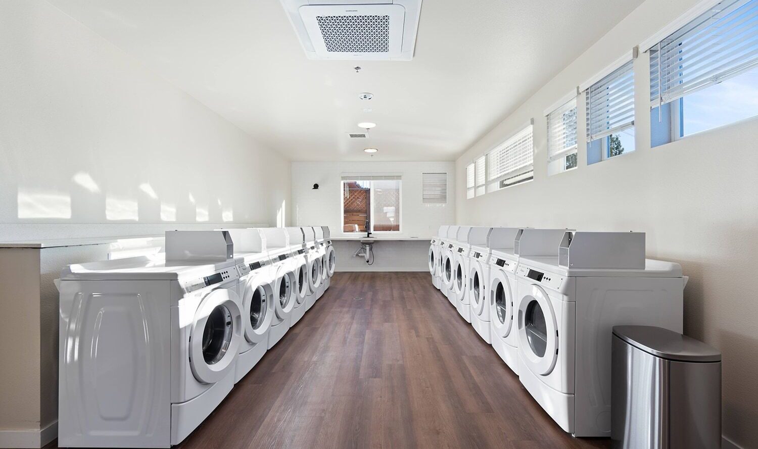 A bright laundry room with two rows of white washing machines and dryers, wooden flooring, and large windows allowing natural light. A trash bin is placed near the machines.