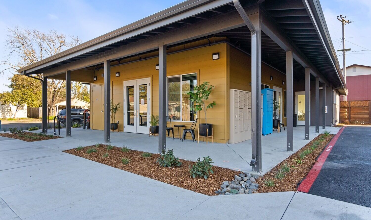 A small modern building with a yellow and gray exterior, featuring a covered porch with seating. There are plants along the walkway, and a bright blue door is visible on the side. A car is parked nearby under a clear blue sky.