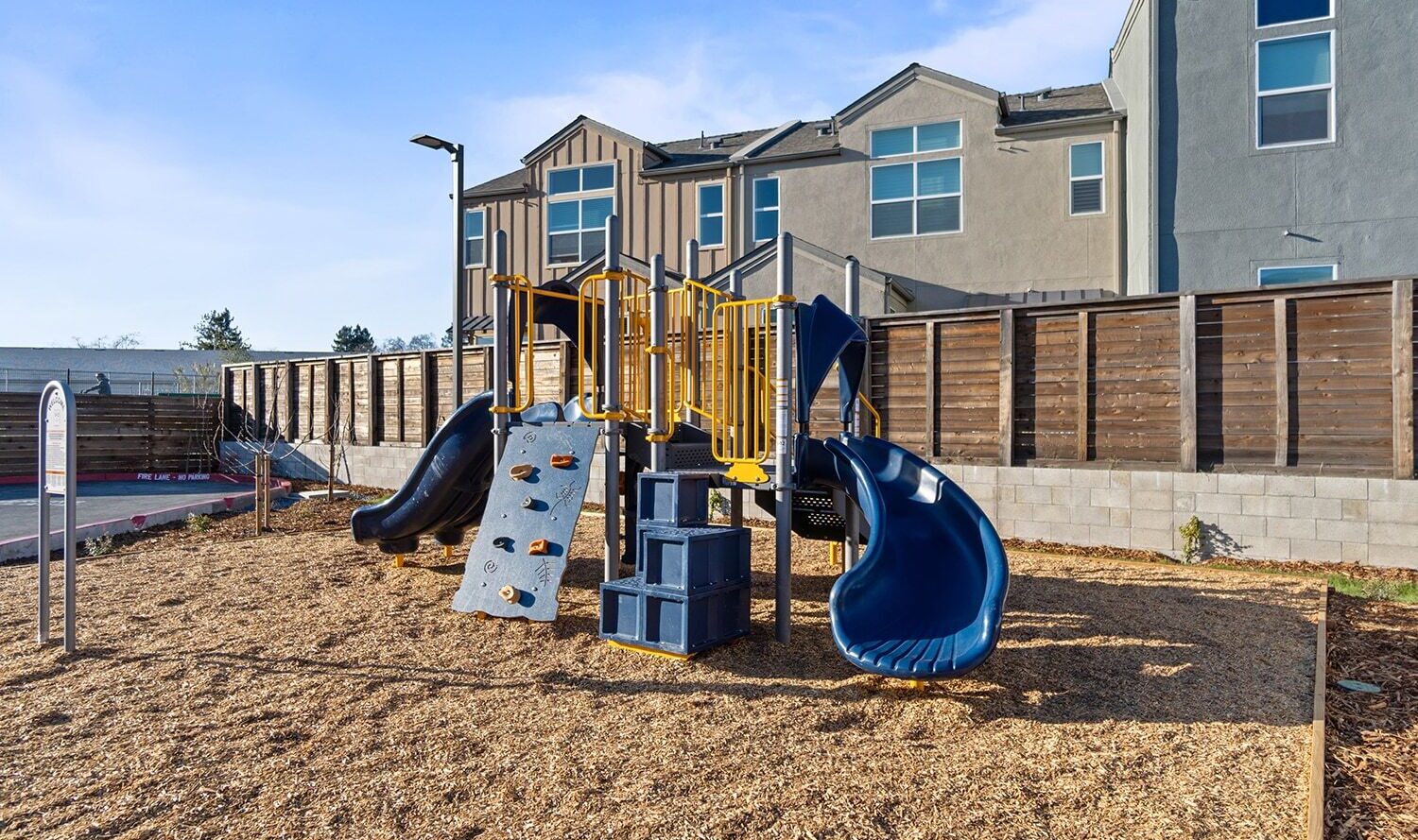 A playground with a climbing wall, steps, and two blue slides on a wood chip surface. The area is fenced with wooden panels and is near residential buildings under a clear blue sky.