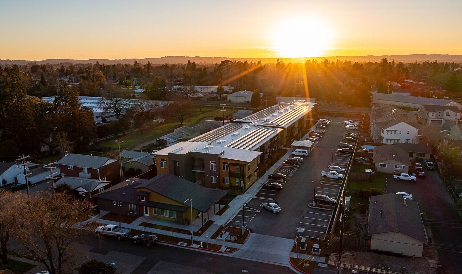 Aerial view of a suburban neighborhood at sunset. A long building with solar panels is in the center, surrounded by parked cars and houses. The sun sets in the horizon, casting a warm glow over the scene.
