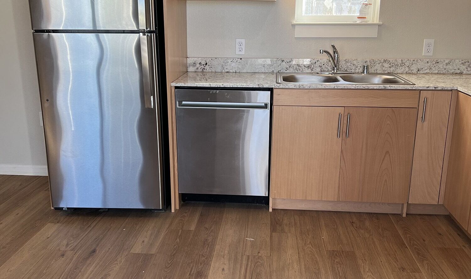Modern kitchen with wooden cabinets, stainless steel refrigerator, dishwasher, and a marble countertop. A window above the sink allows natural light to enter, and there is a wooden floor.