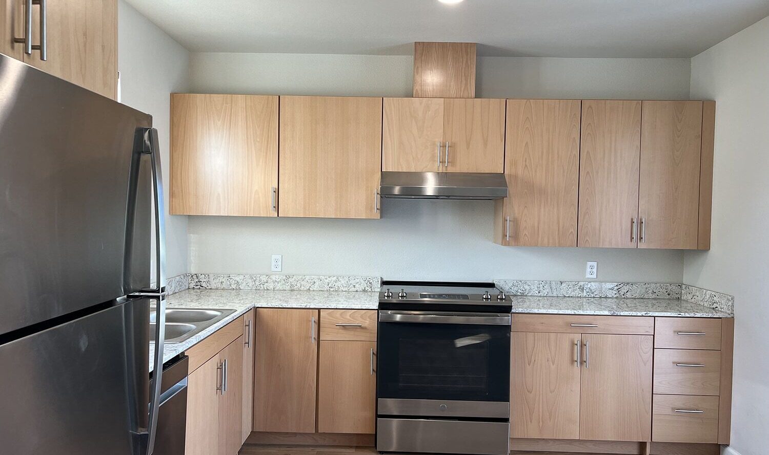 A modern kitchen with light wood cabinets, stainless steel fridge, and a built-in oven. The countertop is a speckled gray and white granite, and the floor is a natural wood finish. A ceiling light is illuminated above.