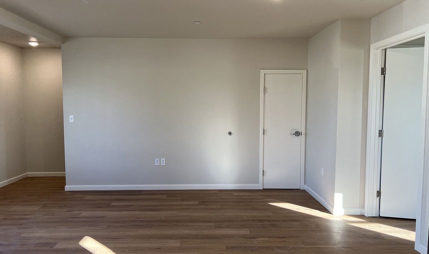 A bright, empty room with light wood flooring and neutral-colored walls. Sunlight streams through a window, casting shadows on the floor. There is a small closed door on the right and an open doorway leading to another room. Ceiling lights are on.