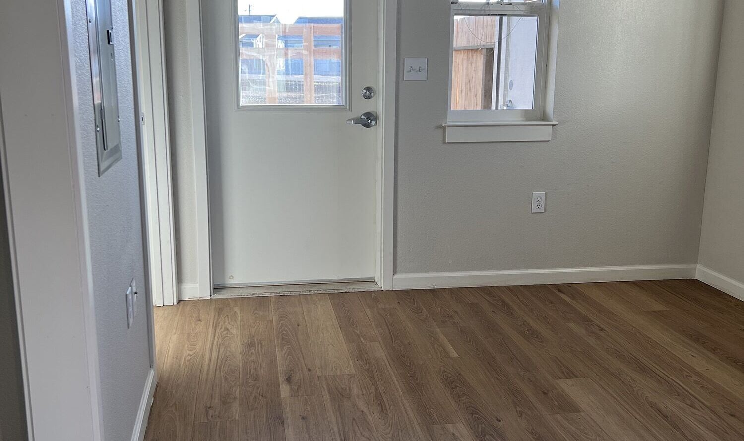 A small room with light-colored walls and wooden floors. A white door with a glass panel leads outside. Theres a window to the right of the door, allowing natural light into the space.
