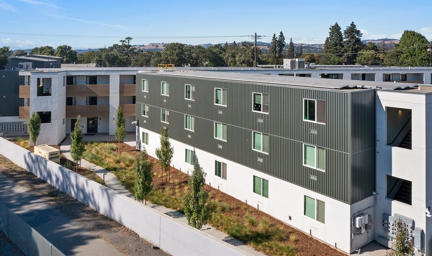 A modern three-story apartment complex with green and white exterior panels. It features several windows, a landscaped area with small trees and shrubs, and a light gray boundary wall. Trees and a blue sky are visible in the background.