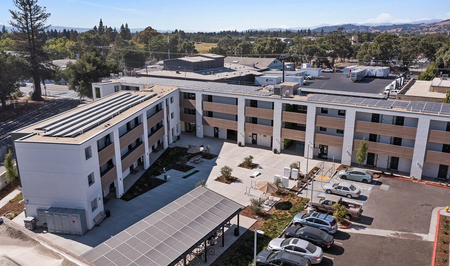 Aerial view of a modern apartment complex with solar panels on the roof. The courtyard features landscaping and bike racks. Several parked cars are seen in the parking lot. The area is surrounded by trees and distant hills under a clear blue sky.