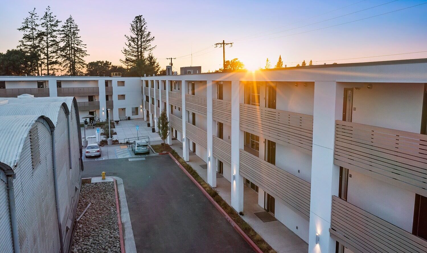 A modern apartment complex with multiple stories and minimalistic balconies, set against a backdrop of tall trees at sunset. A paved driveway leads through the complex, and soft exterior lights illuminate the buildings.