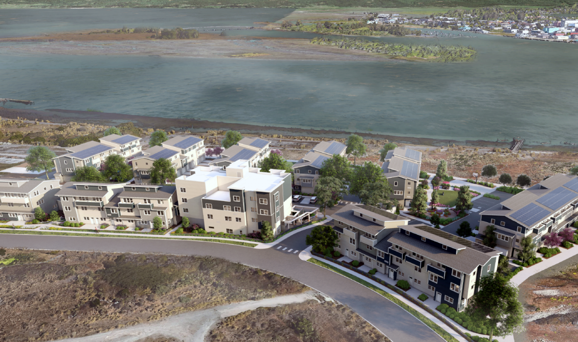 Aerial view of a modern housing complex near a body of water. The complex features multiple multi-story buildings, surrounded by greenery and pathways. In the background, water and distant land areas are visible under a cloudy sky.