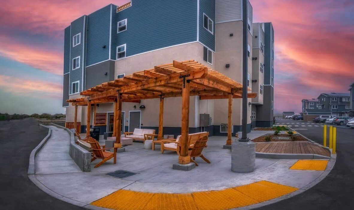 A modern apartment building with a wooden pergola seating area in the foreground. The sky is a vibrant pink and purple at sunset. The scene is surrounded by a parking lot and additional buildings.