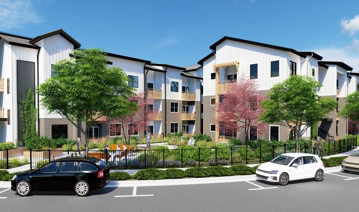 A modern apartment complex with a white facade, surrounded by trees and landscaping. Several cars are parked on the street, and people are walking near a fenced garden area. The sky is clear and blue.