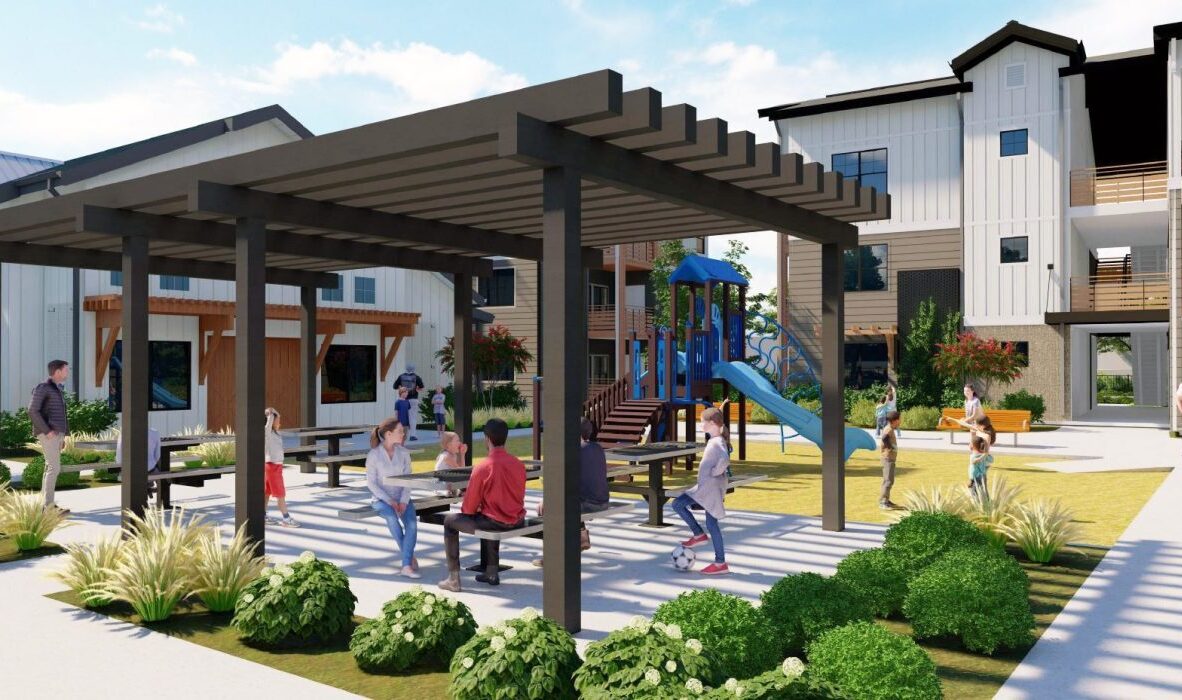 A courtyard with a pergola covering picnic tables. Several people are seated and talking, while children play on a nearby playground with a slide. Modern multi-story buildings surround the area under a clear blue sky.