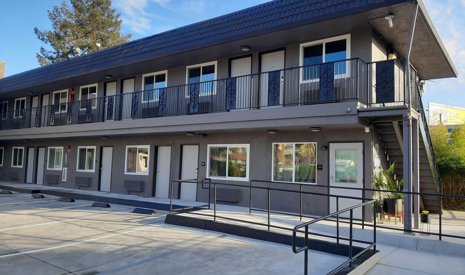 A two-story gray building with a dark roof and black railings. The facade features various windows and doors. A ramp and stairs provide access to the upper level. A tree and clear blue sky are visible in the background.