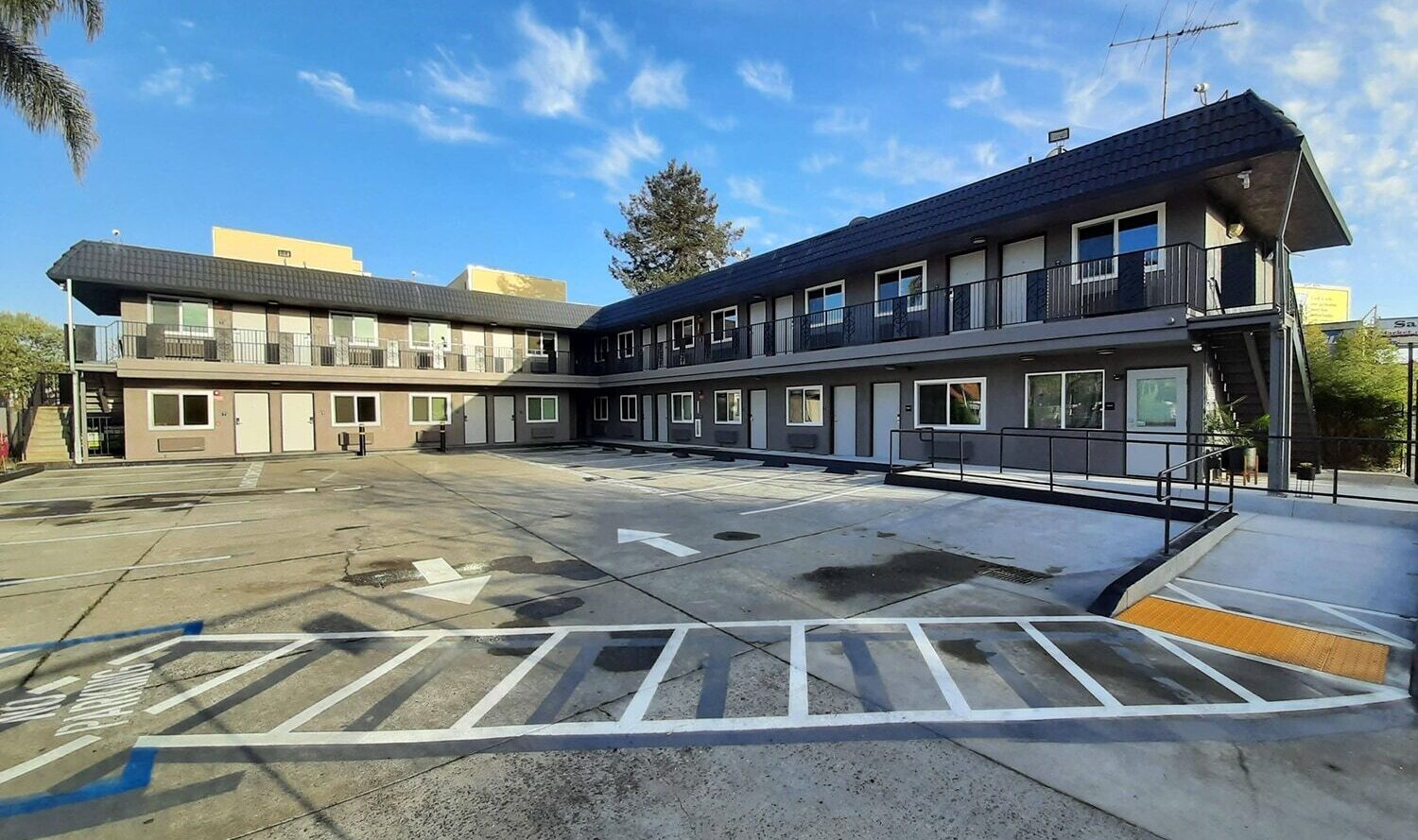 A two-story motel building with a gray exterior and balconies on each floor. The parking area in front has a few marked spaces and accessibility ramps. A clear blue sky with scattered clouds is overhead.
