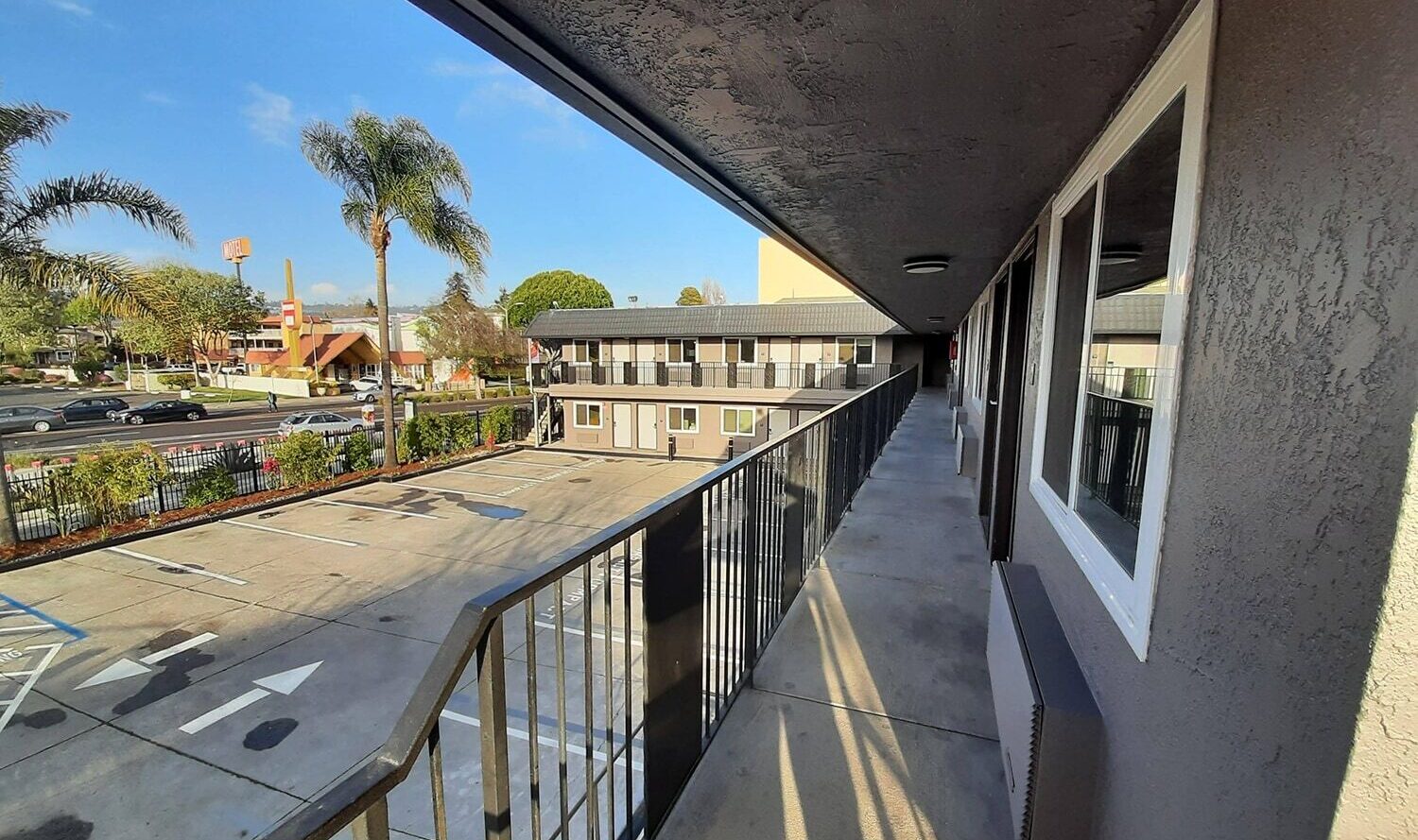 A view from a second-floor walkway of a motel, showing a parking lot with a handicapped space, palm trees, and nearby buildings under a clear blue sky. The walkway has black railings and windows along the wall.