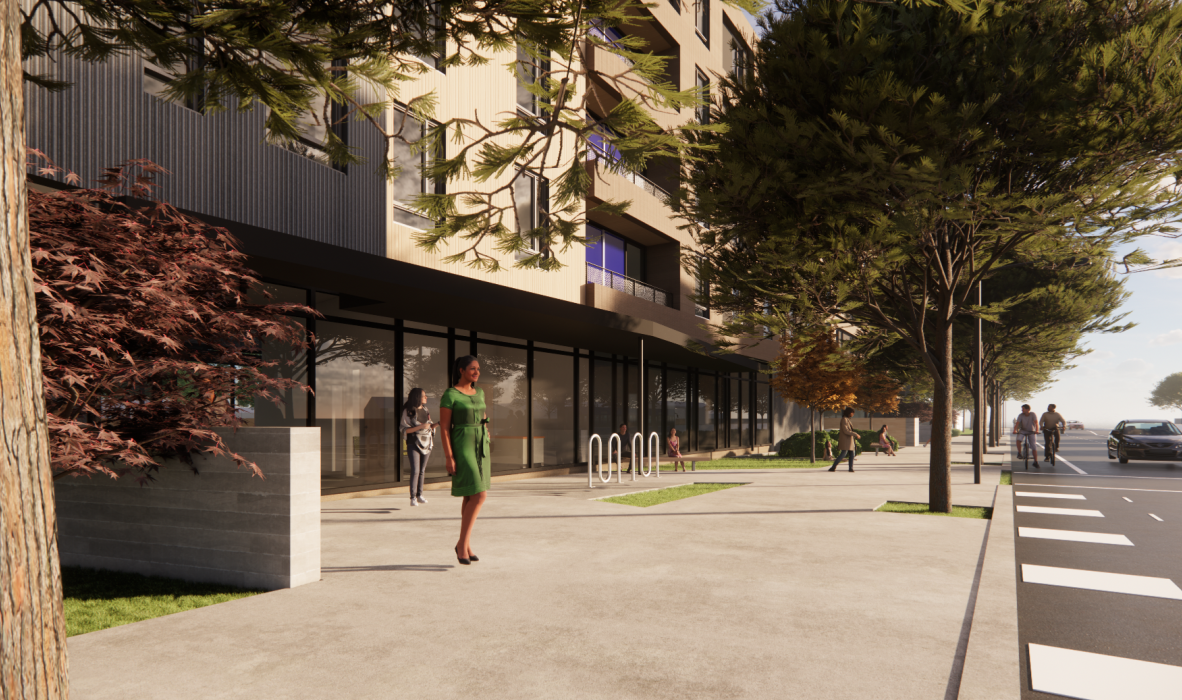 A woman in a green dress walks along a modern, tree-lined sidewalk. The building in the background has large glass windows and a bike rack nearby. Several other people are visible strolling along the path on a sunny day.